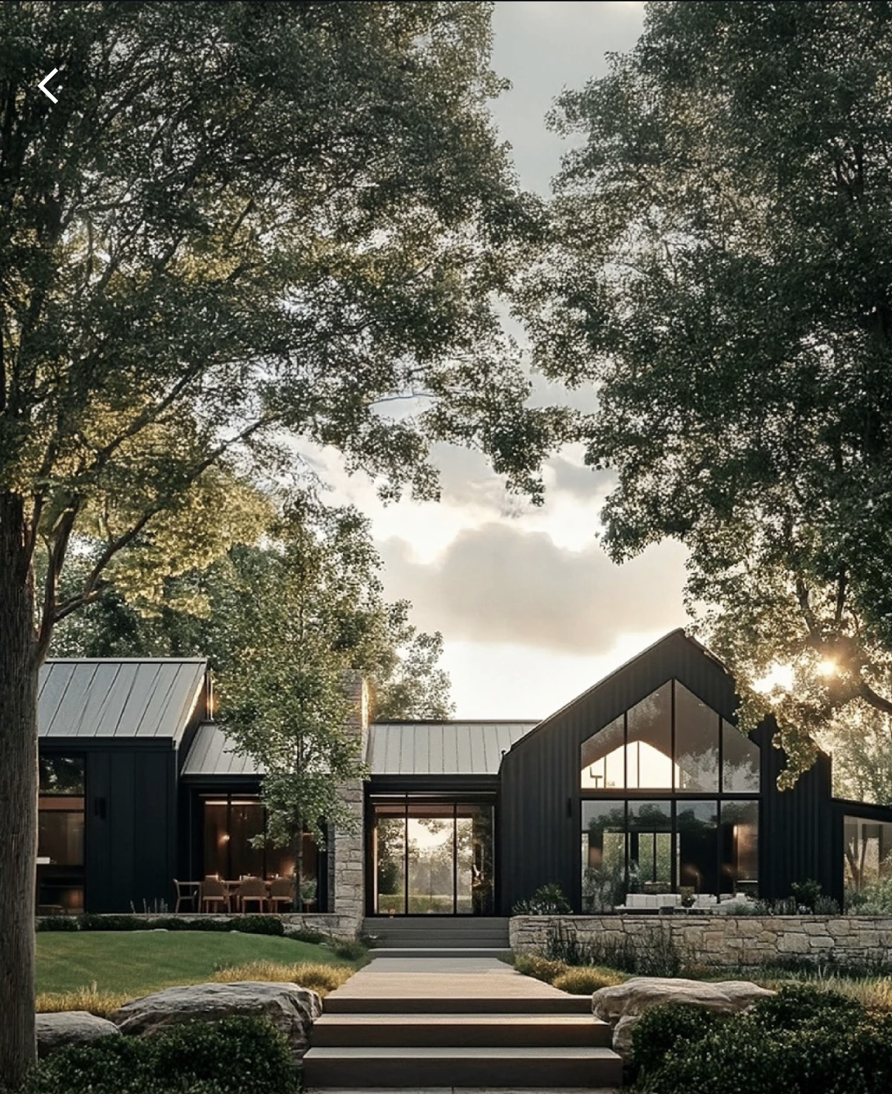 Modern black barndominium with dramatic glass gable end, metal roof, and stone accents framed by mature trees
